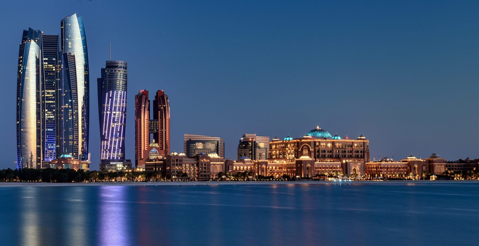 Abu Dhabi skyline at dusk with tall towers and a large domed palace hotel reflected in the water